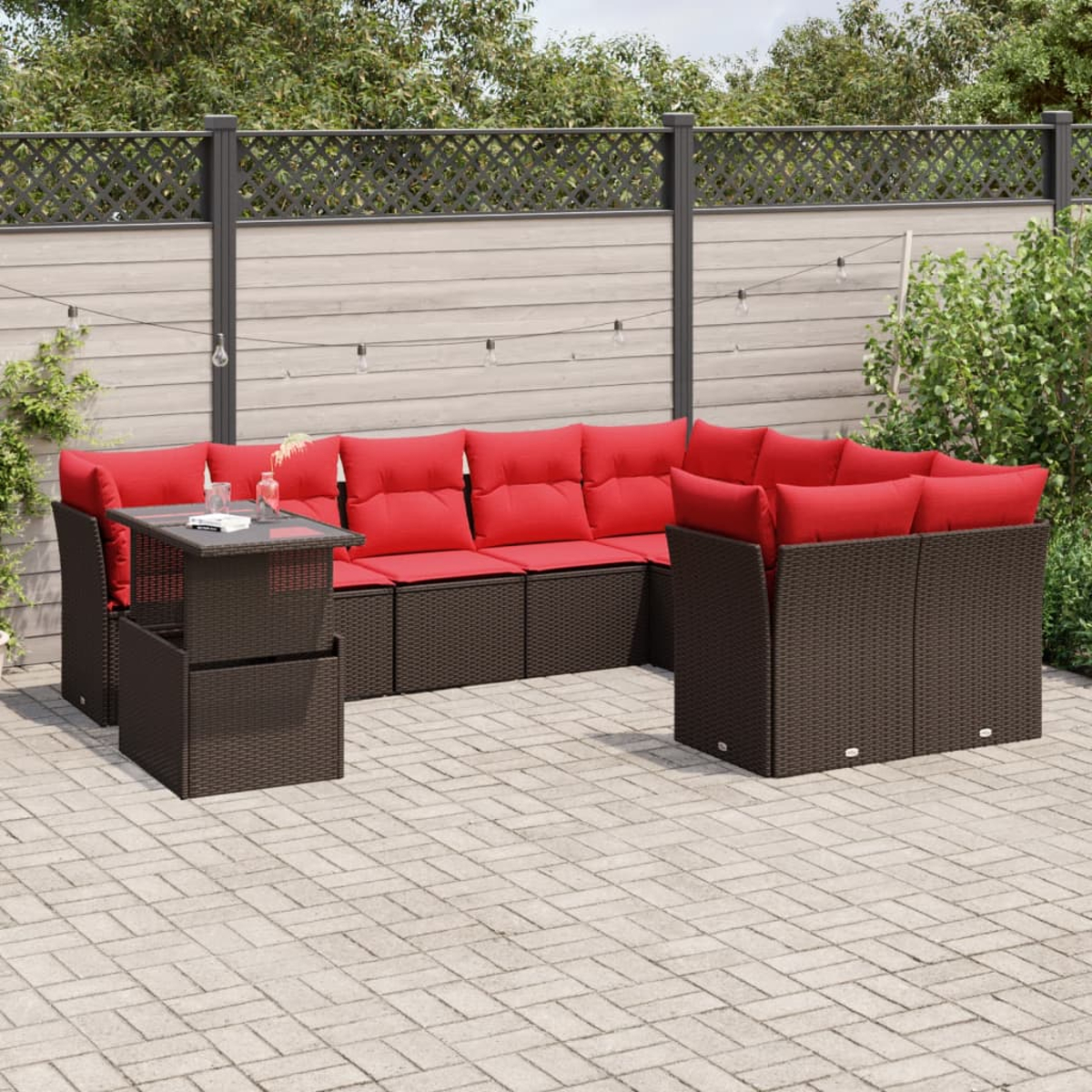 Terrassenset mit roten Kissen. Beinhaltet einen Tisch und ein Ecksofa auf einer Backsteinterrasse. Im Hintergrund sind ein Zaun und Pflanzen zu sehen.