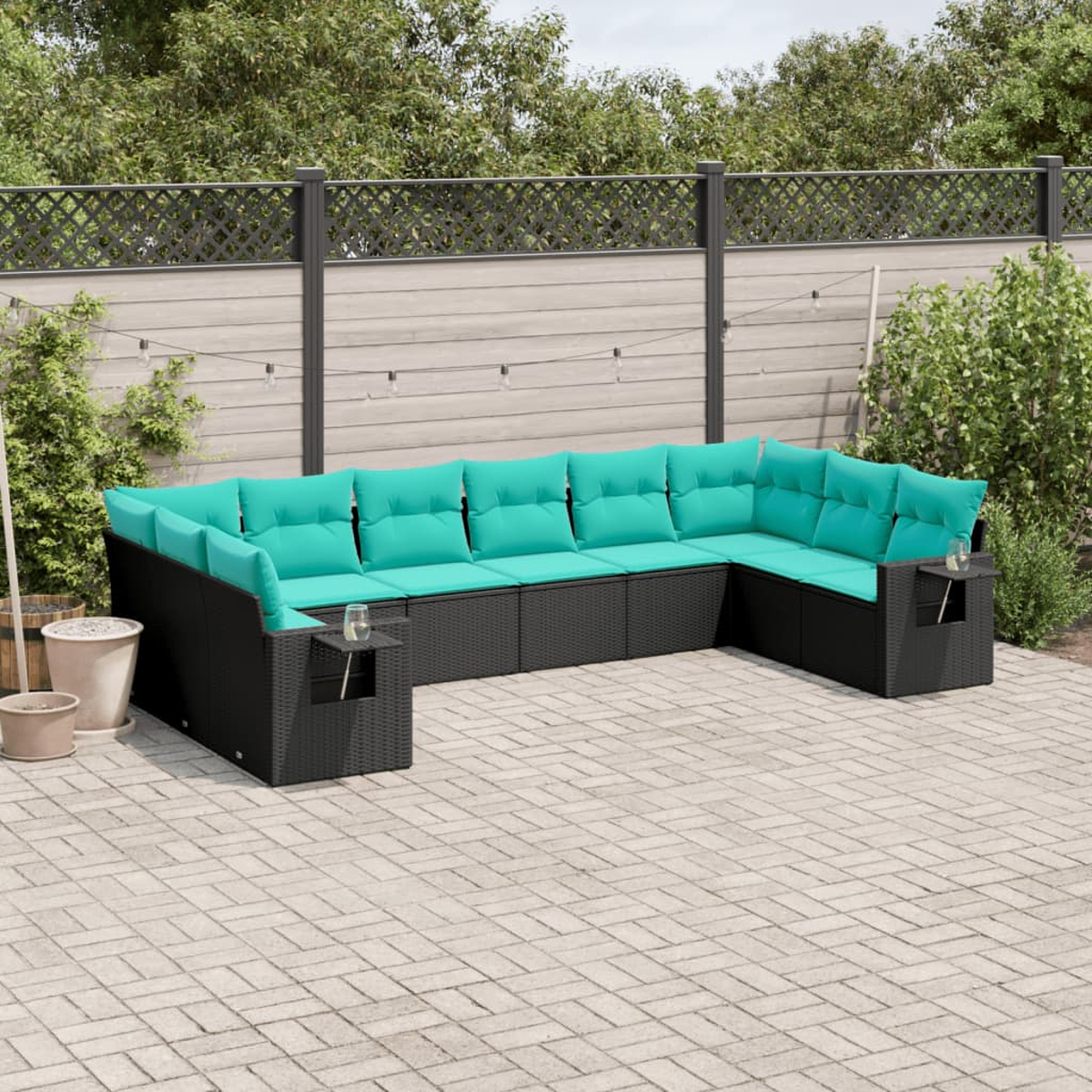 Ein Ecksofa im Freien mit schwarzem, gewebtem Rahmen und türkisfarbenen Kissen steht auf einer Terrasse. Im Hintergrund befinden sich ein Zaun und Grün.