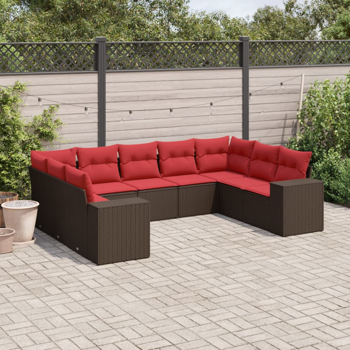 Ein braunes Korbsessel-Sofa mit roten Kissen steht auf einer Terrasse. Dahinter befinden sich ein Zaun und Grün. Lichterketten hängen oben.