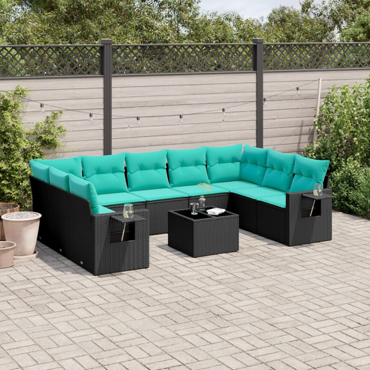 Schwarzes Outdoor-Ecksofa aus Korbgeflecht mit türkisfarbenen Kissen und einem kleinen Tisch auf einer Terrasse. Hintergrund mit Zaun und Grün.