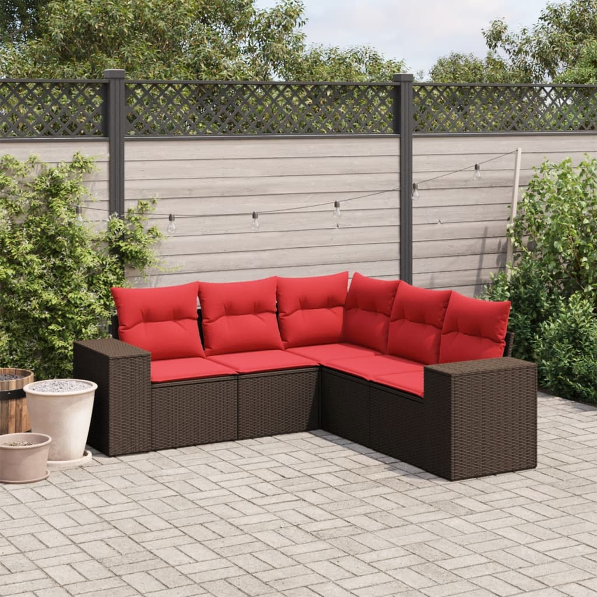 Braunes Ecksofa aus Weide für den Außenbereich mit roten Kissen auf einer gepflasterten Terrasse. Im Hintergrund ein Holzzaun und Pflanzen.