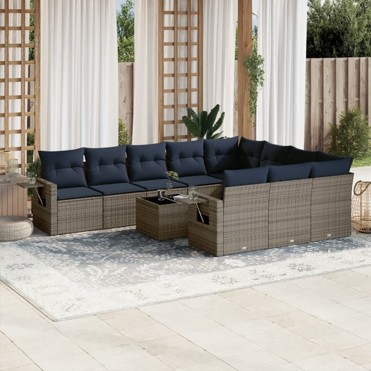 Outdoor-Ecksofa mit marineblauen Kissen auf einem grauen Teppich. Weiße Vorhänge und Spalier, Pflanzen im Hintergrund und ein Holzzaun.