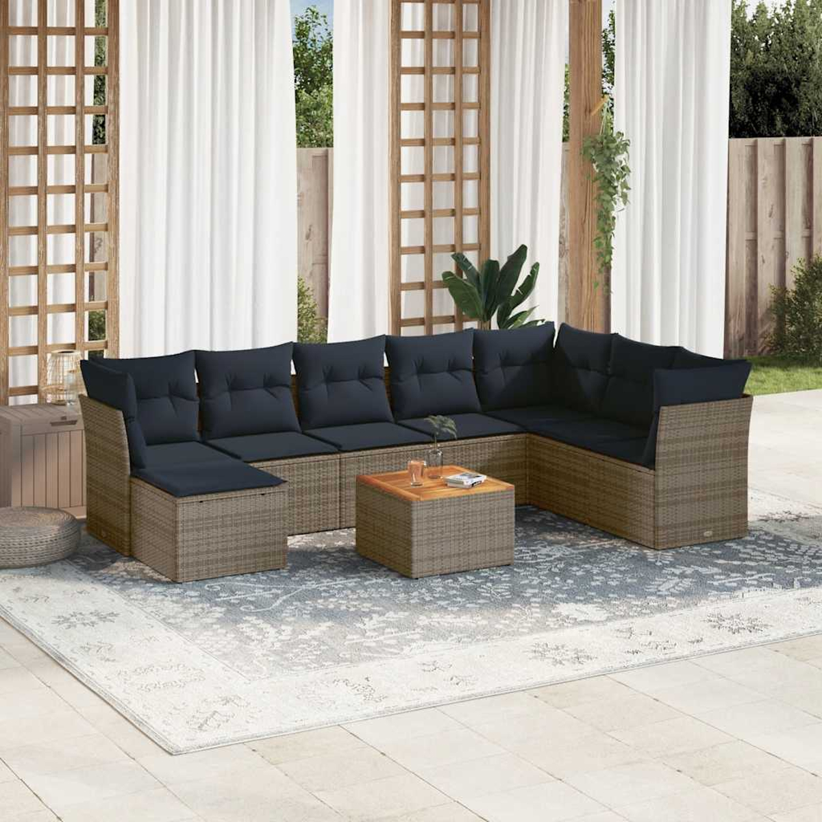 Zona de estar al aire libre con un sofá de mimbre, cojines azul oscuro y una mesa de centro de madera sobre una alfombra estampada. Cortinas blancas al fondo.