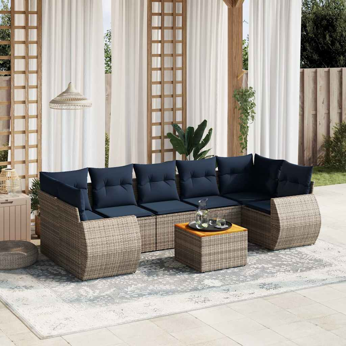 Outdoor-Sitzbereich mit Rattansofa, blauen Kissen und Holztisch. Weiße Vorhänge und Teppich, Grün im Hintergrund.