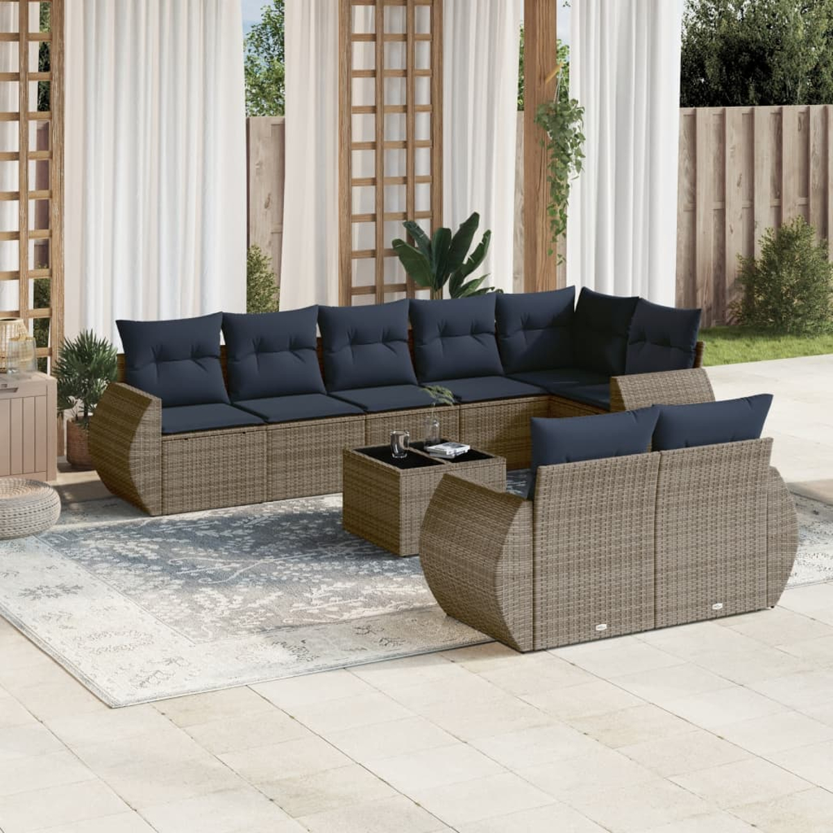 Terrassenset mit gewebten braunen Sitzen, dunkelblauen Kissen und einem quadratischen Couchtisch. Weiße Vorhänge und Grün im Hintergrund.