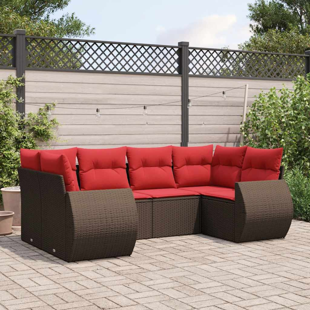 Outdoor-Ecksofa mit roten Kissen. Brauner Korbrahmen auf einer Ziegelterrasse. Zaun und Grün im Hintergrund.