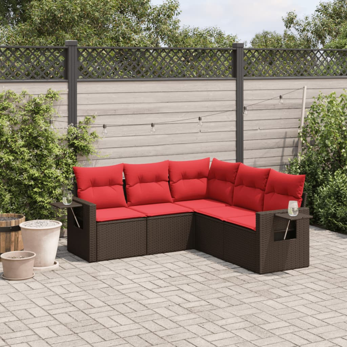 Outdoor-Ecksofa mit roten Kissen. Brauner Korbrahmen. Eine Steinterrasse, Pflanzen und ein Holzzaun befinden sich im Hintergrund.
