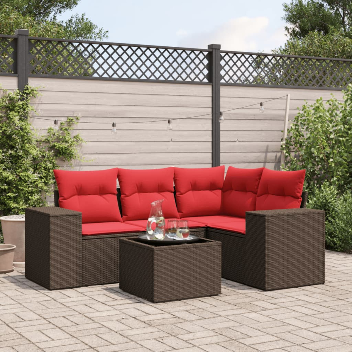 Outdoor-Ecksofa mit roten Kissen, braunen Korbmöbeln, einem Glastisch mit Getränken und einem Zaun im Hintergrund.