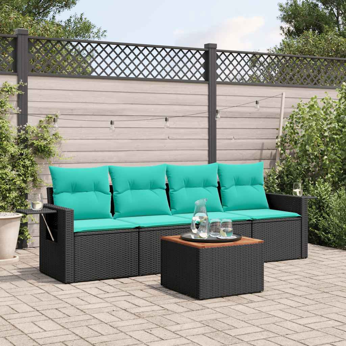 Schwarzes Rattansofa mit türkisfarbenen Kissen und einem Couchtisch auf einer Terrasse. Im Hintergrund ein Zaun und Grün.