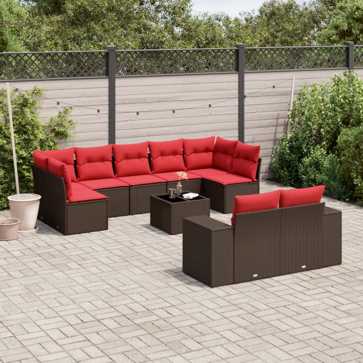 Juego de muebles de patio. Sofás de mimbre marrón con cojines rojos. Mesa pequeña al frente. Patio de piedra gris, vegetación detrás.