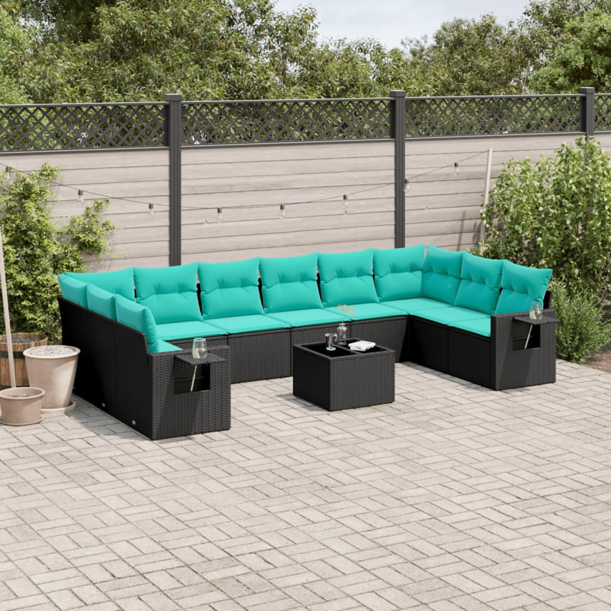 Gartenmöbel mit schwarzem Rattangeflecht, türkisfarbenen Kissen und einem kleinen Tisch. Auf einer Terrasse, grünes Laub im Hintergrund.