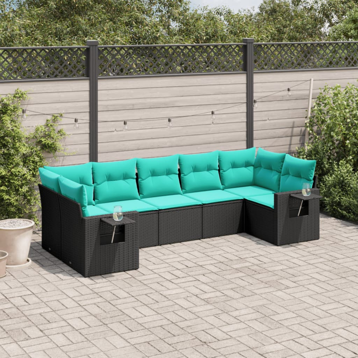 Ein schwarzes Outdoor-Ecksofa aus Korbgeflecht mit türkisfarbenen Kissen steht auf einer Backstein-Terrasse. Im Hintergrund ein Holzzaun und Grün.
