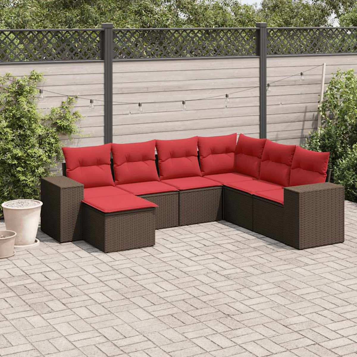 Ein Ecksofa mit roten Kissen steht auf einer Terrasse. Im Hintergrund sind ein Zaun und Grün. Lichterketten hängen darüber.