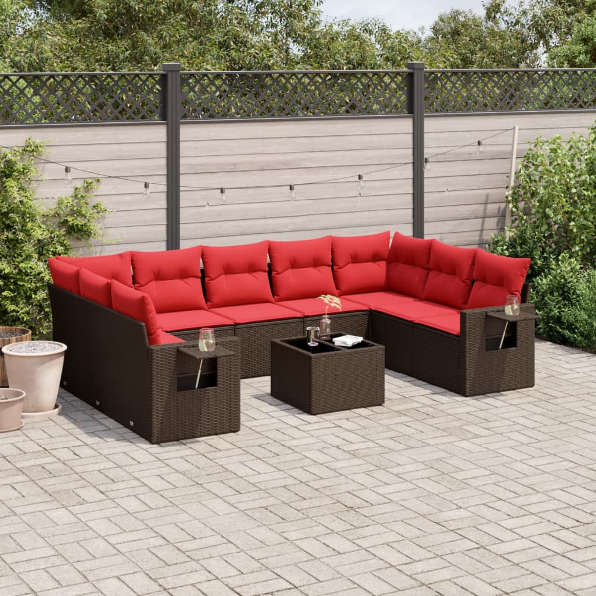 Ecksofa für draußen mit roten Kissen, einem Couchtisch und Beistelltischen. Auf einer gepflasterten Terrasse, mit Zaun und Pflanzen im Hintergrund.