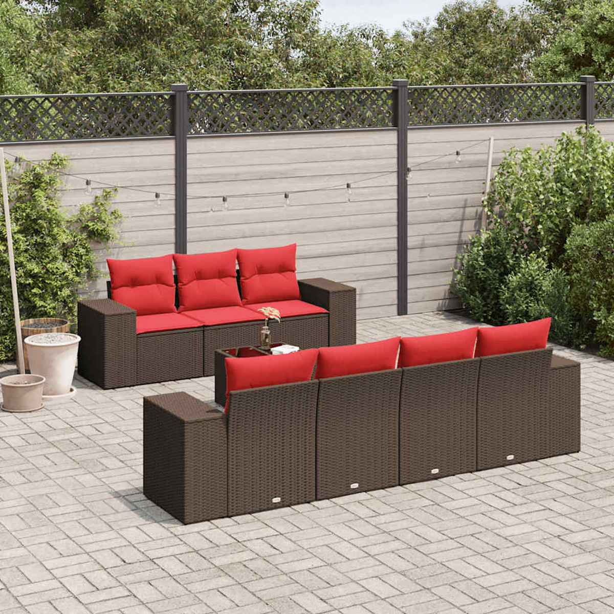 Terrassenmöbel-Set mit braunen Korbs Sofas und roten Kissen. Auf einer Backstein-Terrasse, mit neutralem Zaun und grünem Laub im Hintergrund.