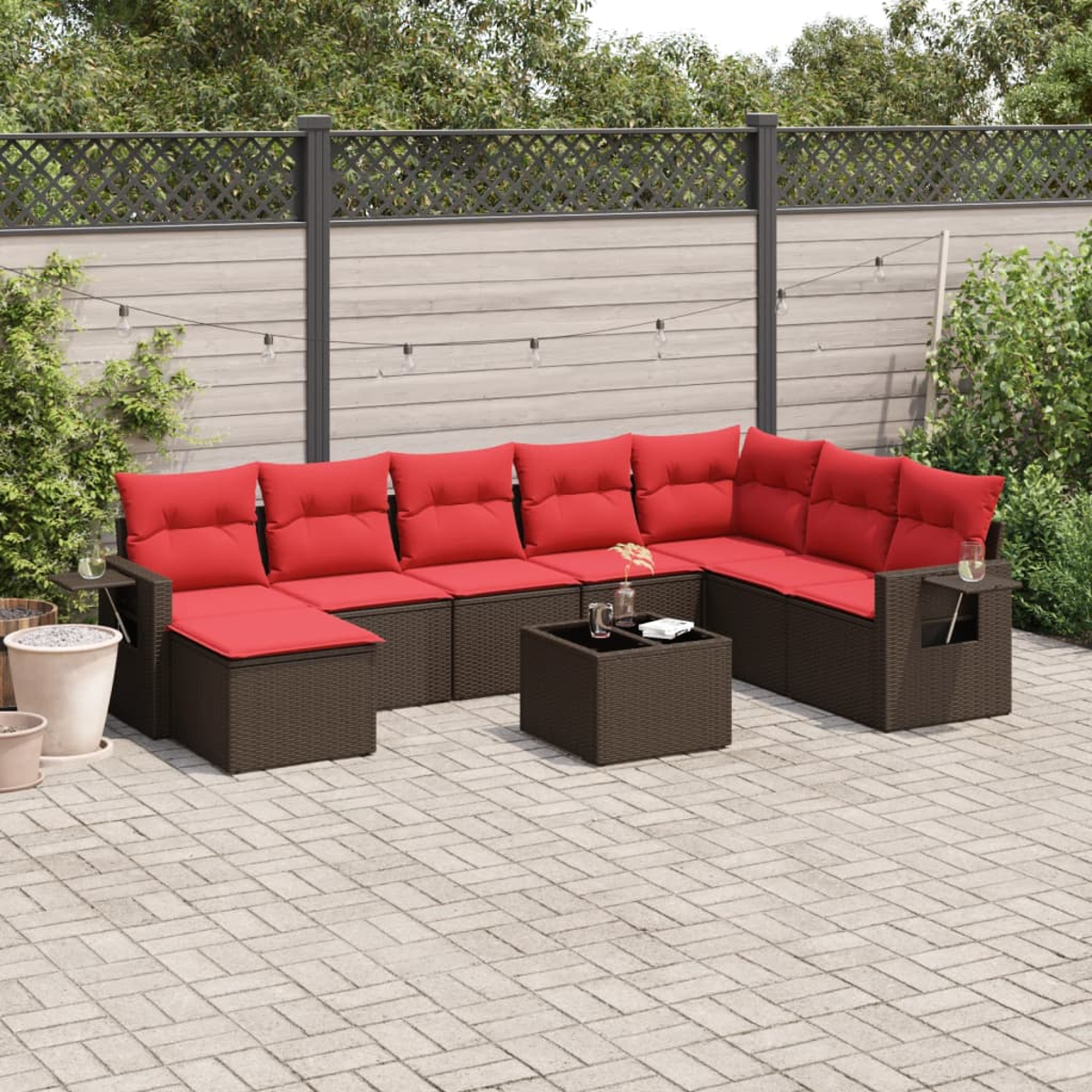Outdoor-Ecksofa mit roten Kissen. Brauner Korbrahmen, Couchtisch und Beistelltische. Auf einer Terrasse mit Zaun und Pflanzen.