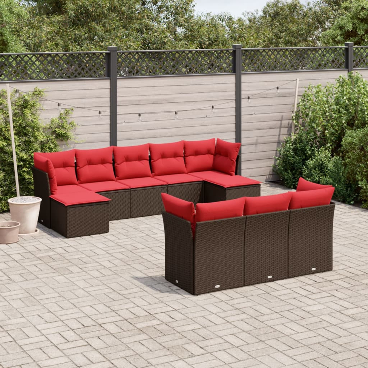 Gartenmöbel-Set mit roten Kissen und braunen Korbflechtrahmen auf einer Terrasse. Hintergrund: Zaun, Grün und Lichterketten.