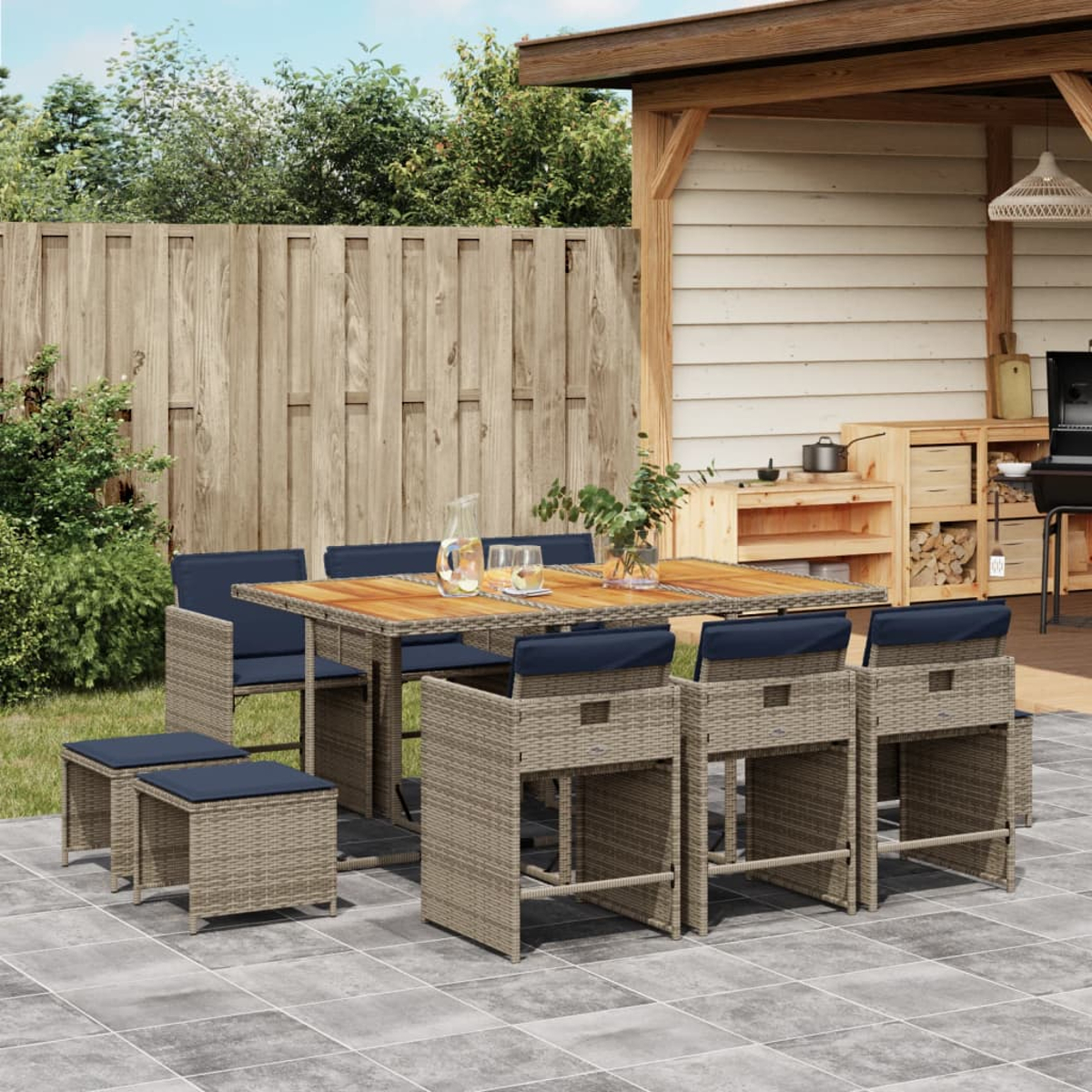 Essgruppe im Freien mit Tisch, Stühlen und Hockern auf einer Terrasse. Holzzaun und -struktur im Hintergrund. Getränke stehen auf dem Tisch.