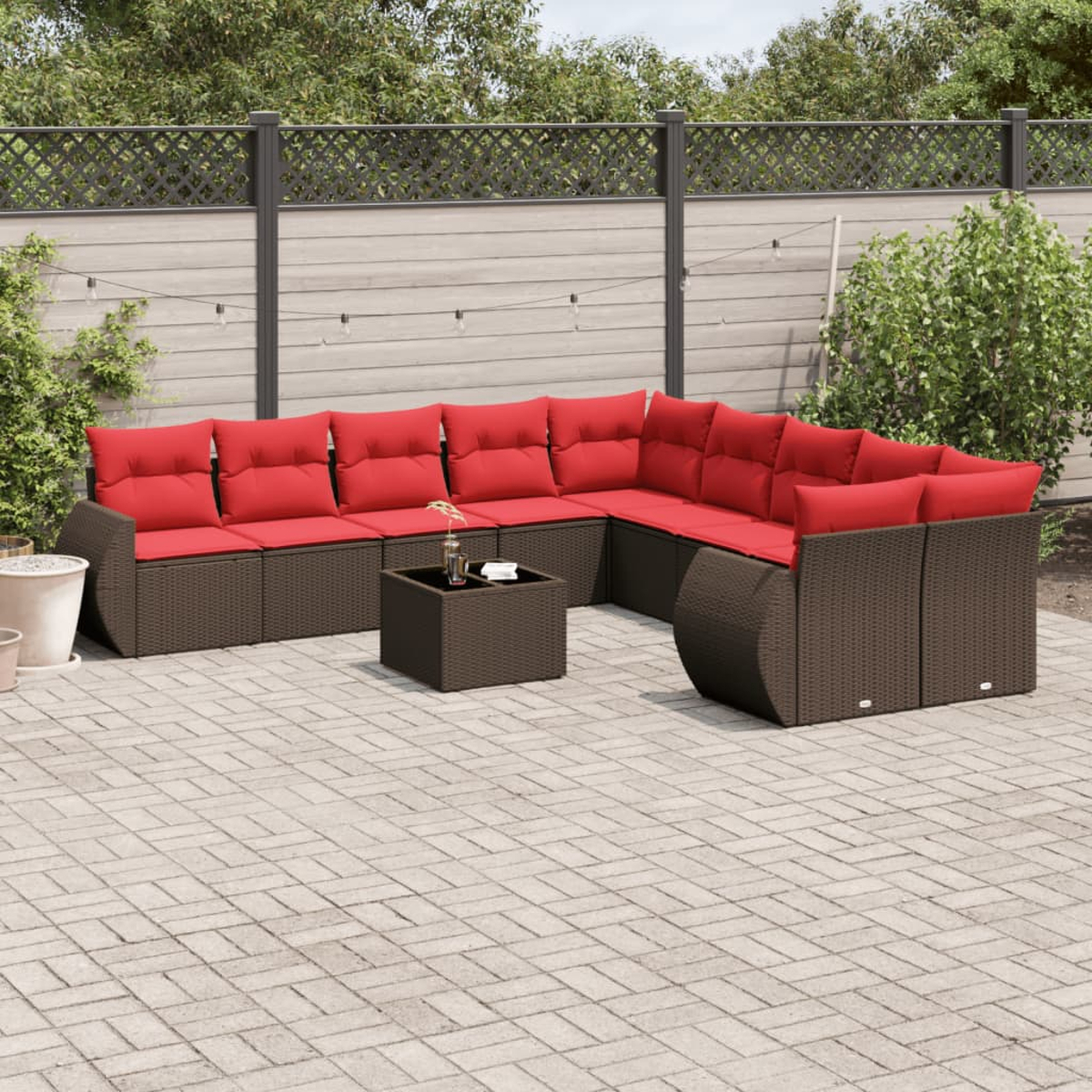 Ein Ecksofa im Freien mit roten Kissen steht auf einer Terrasse. Davor steht ein kleiner Tisch. Im Hintergrund sind ein Zaun und Grün.