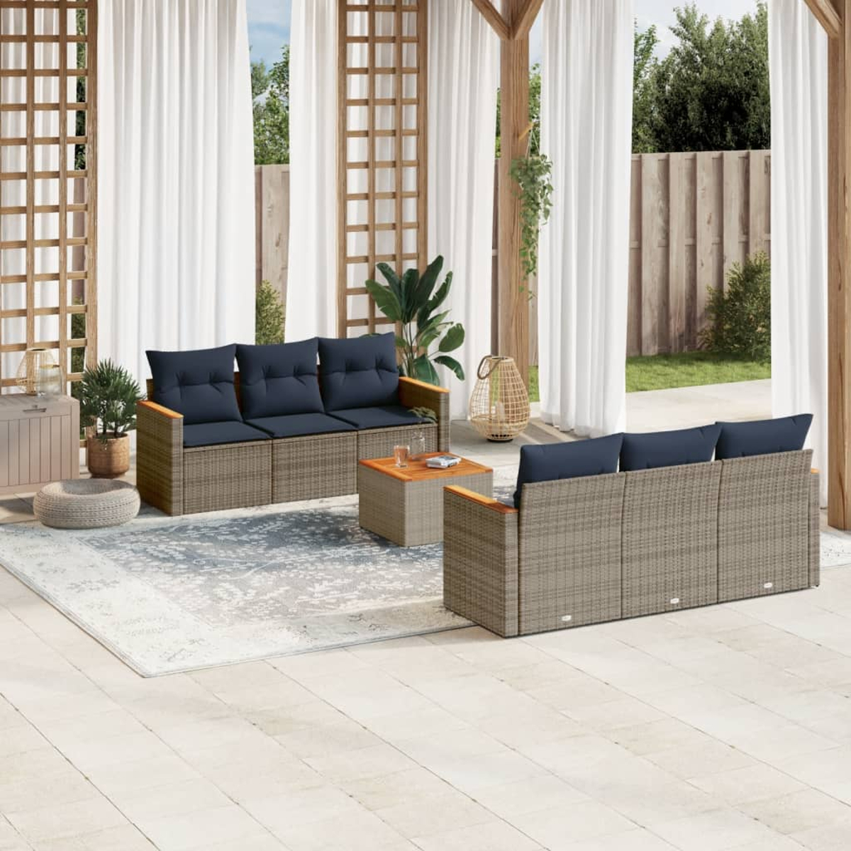 Terrassengarnitur mit grauen Sofas, blauen Kissen, Holztisch und Teppich auf heller Terrasse, mit Vorhängen und Bäumen.