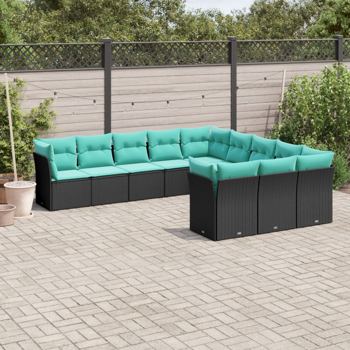 Sofá seccional para exteriores con mimbre negro y cojines turquesa en un patio de ladrillos. Una valla de madera y vegetación al fondo.