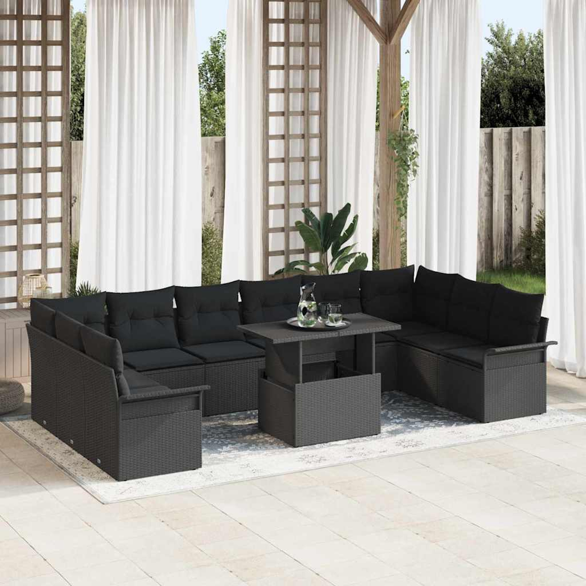 Graues Outdoor-Korbsessel-Sofa mit schwarzen Kissen, auf einem Teppich, mit einem Tisch mit Hebefunktion. Weiße Vorhänge und hölzerne Pergola im Hintergrund.