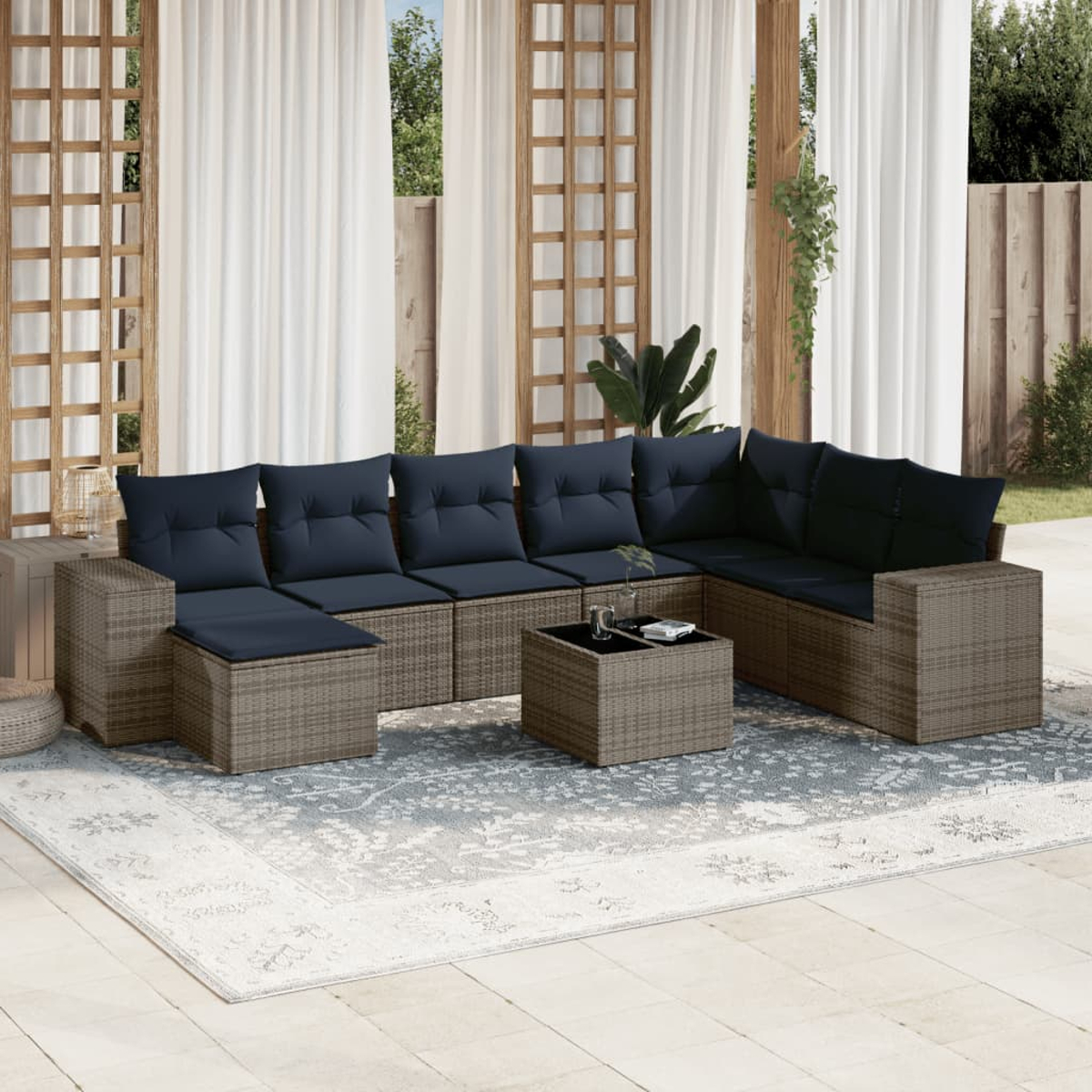 Ein Sitzbereich im Freien mit einem grauen Korbmöbel mit blauen Kissen, einem Couchtisch, einem Teppich und weißen Vorhängen.
