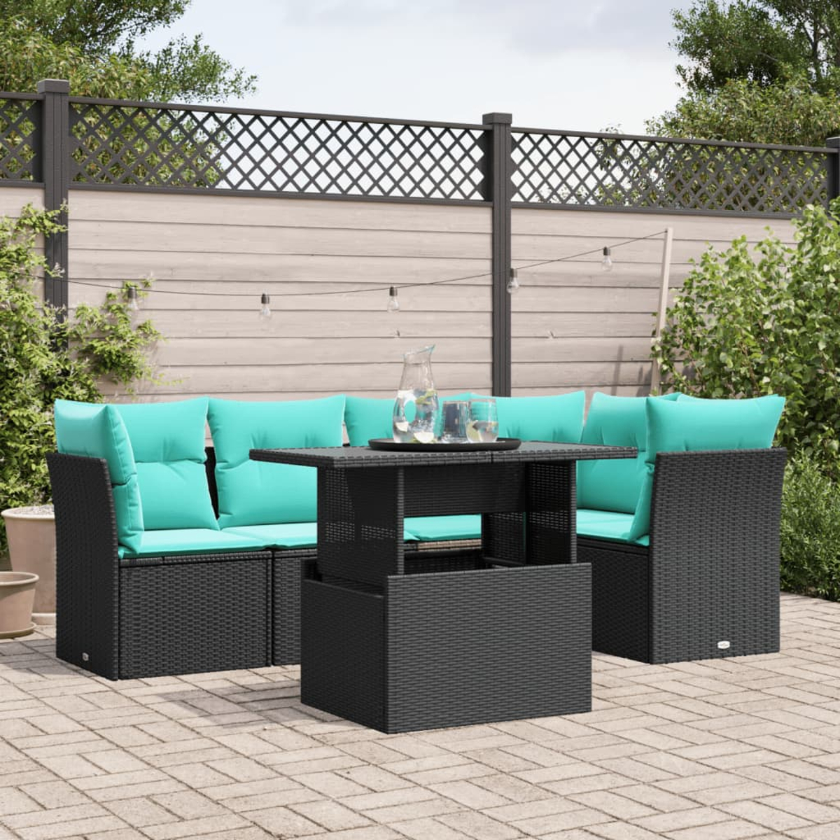 Terrassengarnitur mit schwarzem Korbmöbel und türkisfarbenen Kissen. Ein Tisch mit Glaskrug, Gläsern und einem dekorativen Zaun ist sichtbar.