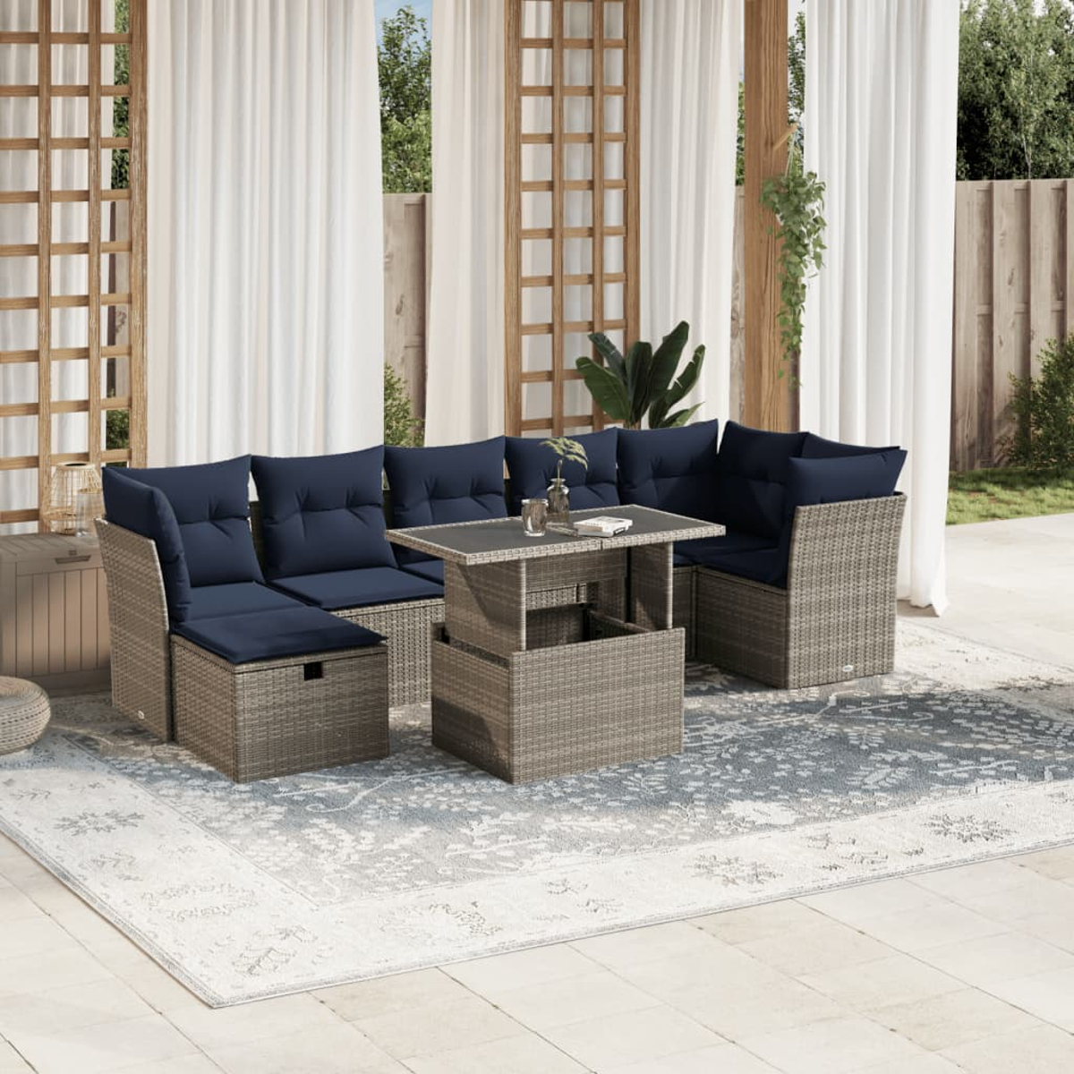 Outdoor-Terrassenset mit blauen Kissen, Korbmöbeln und einem Tisch mit Hubfunktion auf einem gemusterten Teppich, vor weißen Vorhängen.