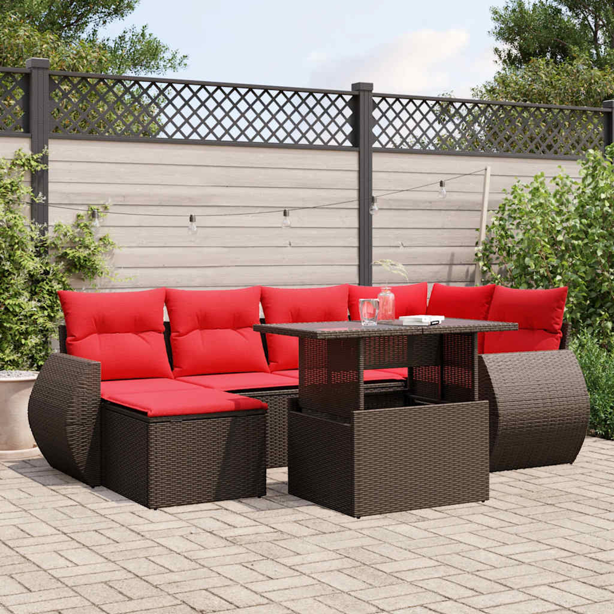 Juego de patio al aire libre con cojines rojos, muebles de mimbre marrón y una mesa de centro. Contra una valla con plantas y un patio de ladrillos.