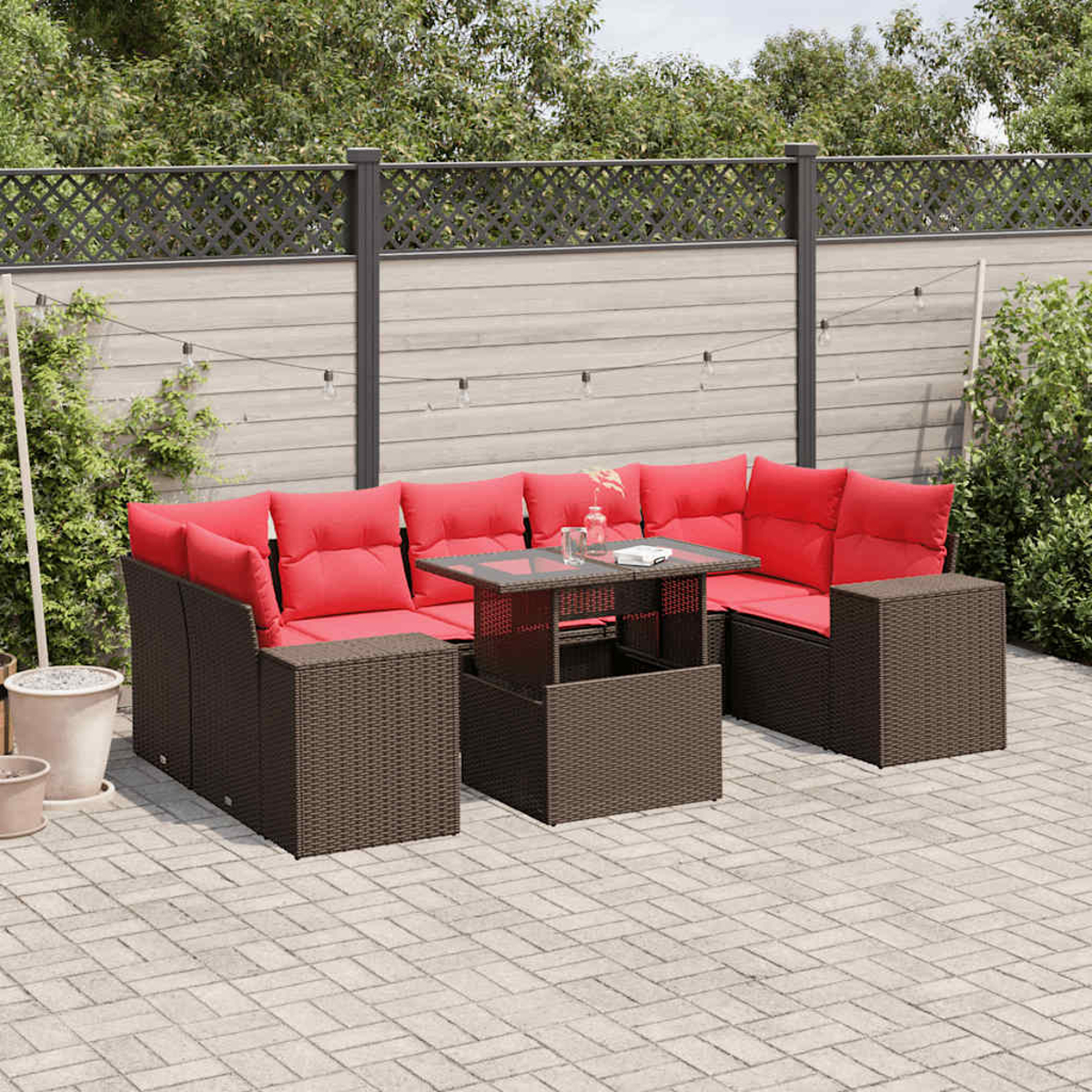 Garten-Sitzgruppe mit roten Kissen und einem Tisch mit Glasplatte auf einer gepflasterten Terrasse, Zaun und Grün im Hintergrund.
