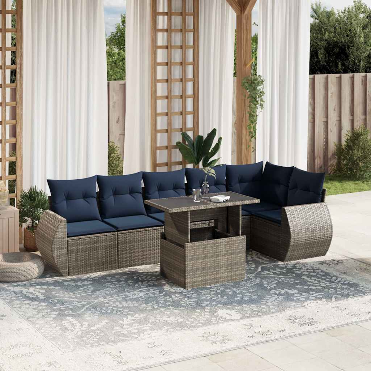 Gartenmöbel mit blauen Kissen, einem grauen Tisch und einem gemusterten Teppich, vor weißen Vorhängen und Grün.