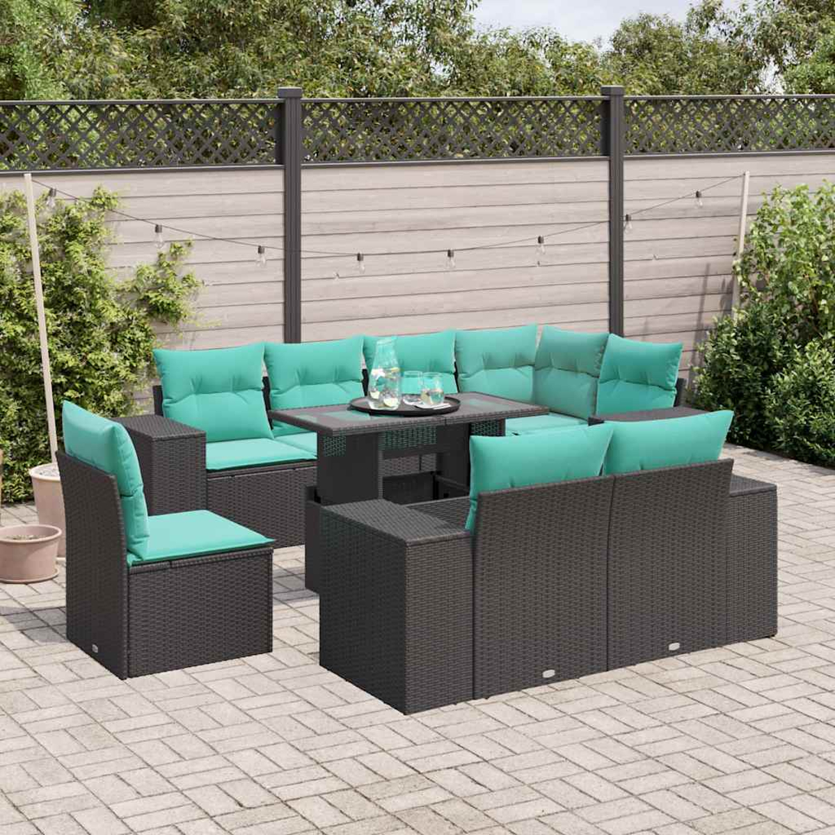 Terrassenmöbel-Set mit grauem Korbmöbeln, türkisfarbenen Kissen, einem kleinen Tisch und einer Ziegelterrasse. Hintergrund: Zaun und Grün.