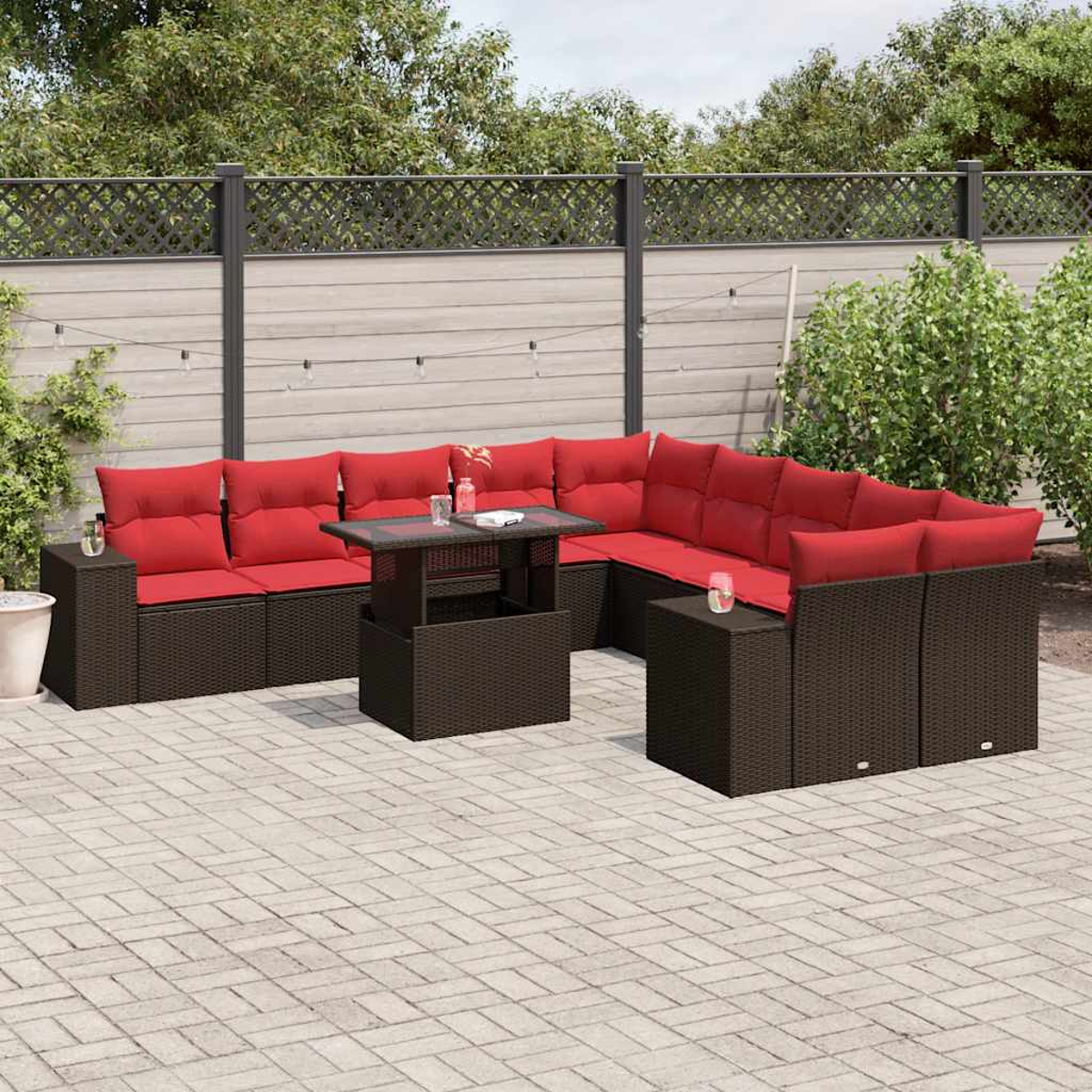 Outdoor-Ecksofa mit roten Kissen und Glastisch. Braune Korbmöbel auf einer Ziegelterrasse, mit Zaun und Grün im Hintergrund.