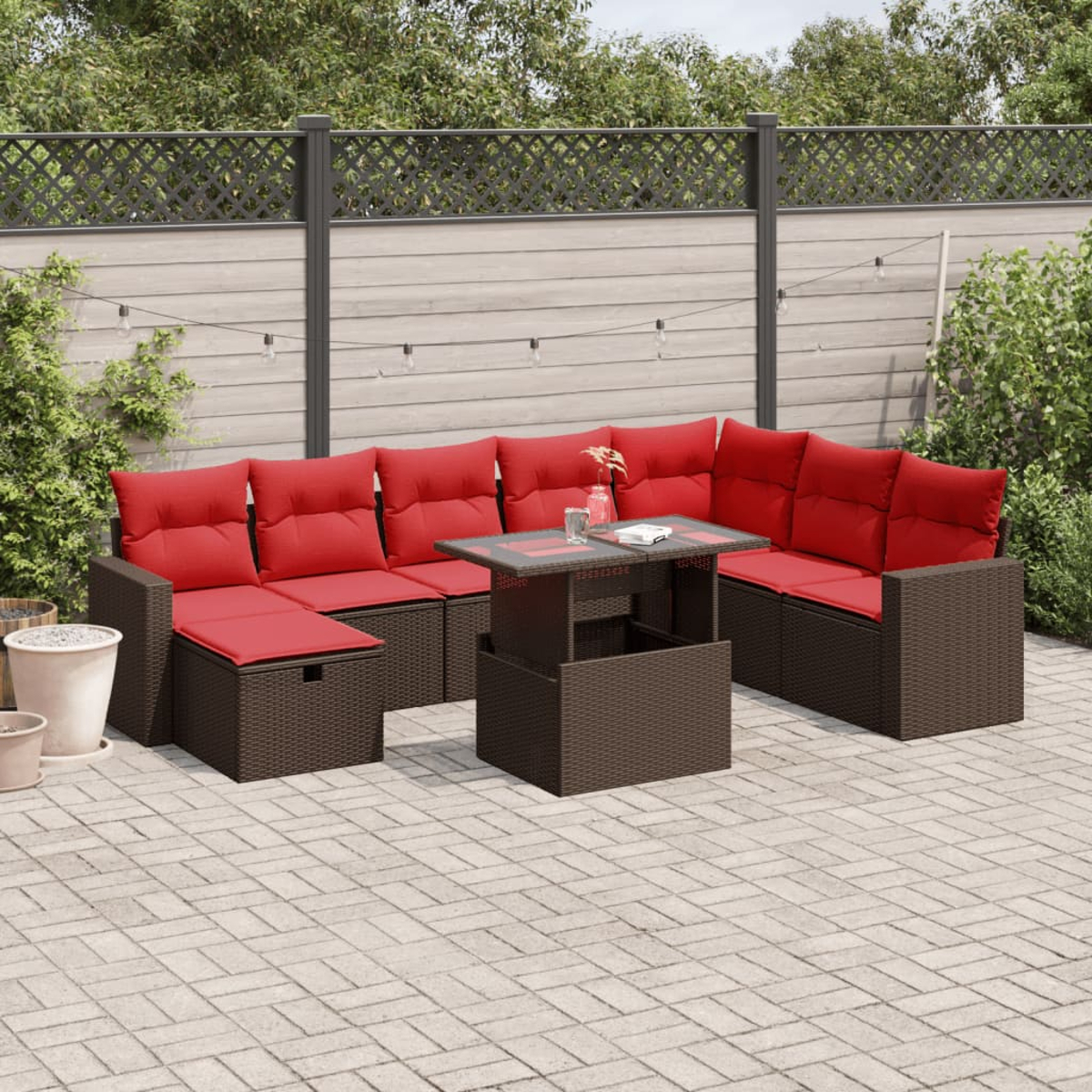 Braune Outdoor-Korbsessel mit roten Kissen und einem Tisch mit Glasplatte auf einer Terrasse. Ein Holzzaun und Pflanzen bilden den Hintergrund.