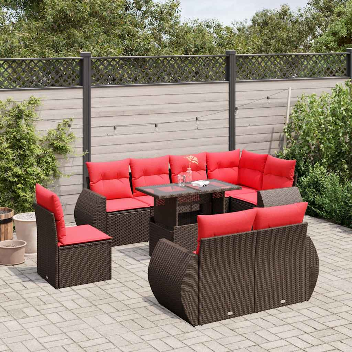 Gartenmöbel-Set. Braune Korbsessel und -sofas mit roten Kissen. Tisch mit Glasplatte. Auf gepflasterter Terrasse. Zaun und Grün im Hintergrund.