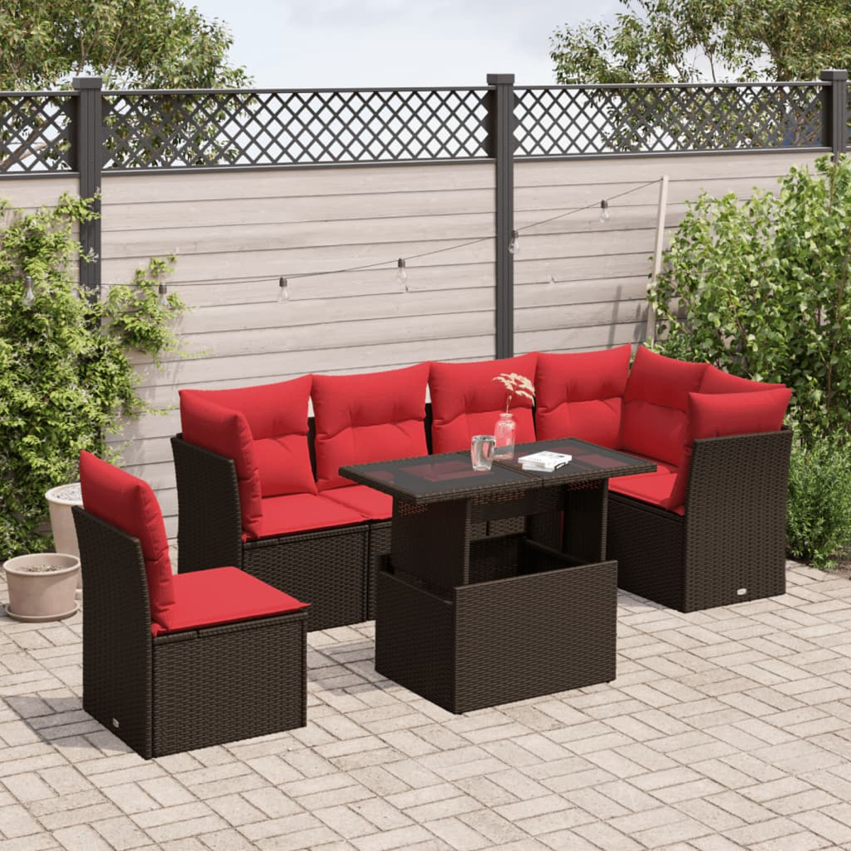 Garten-Set mit roten Kissen, braunem Korbgeflecht und Glastisch. Auf gepflasterter Terrasse mit Zaun und Grün im Hintergrund.