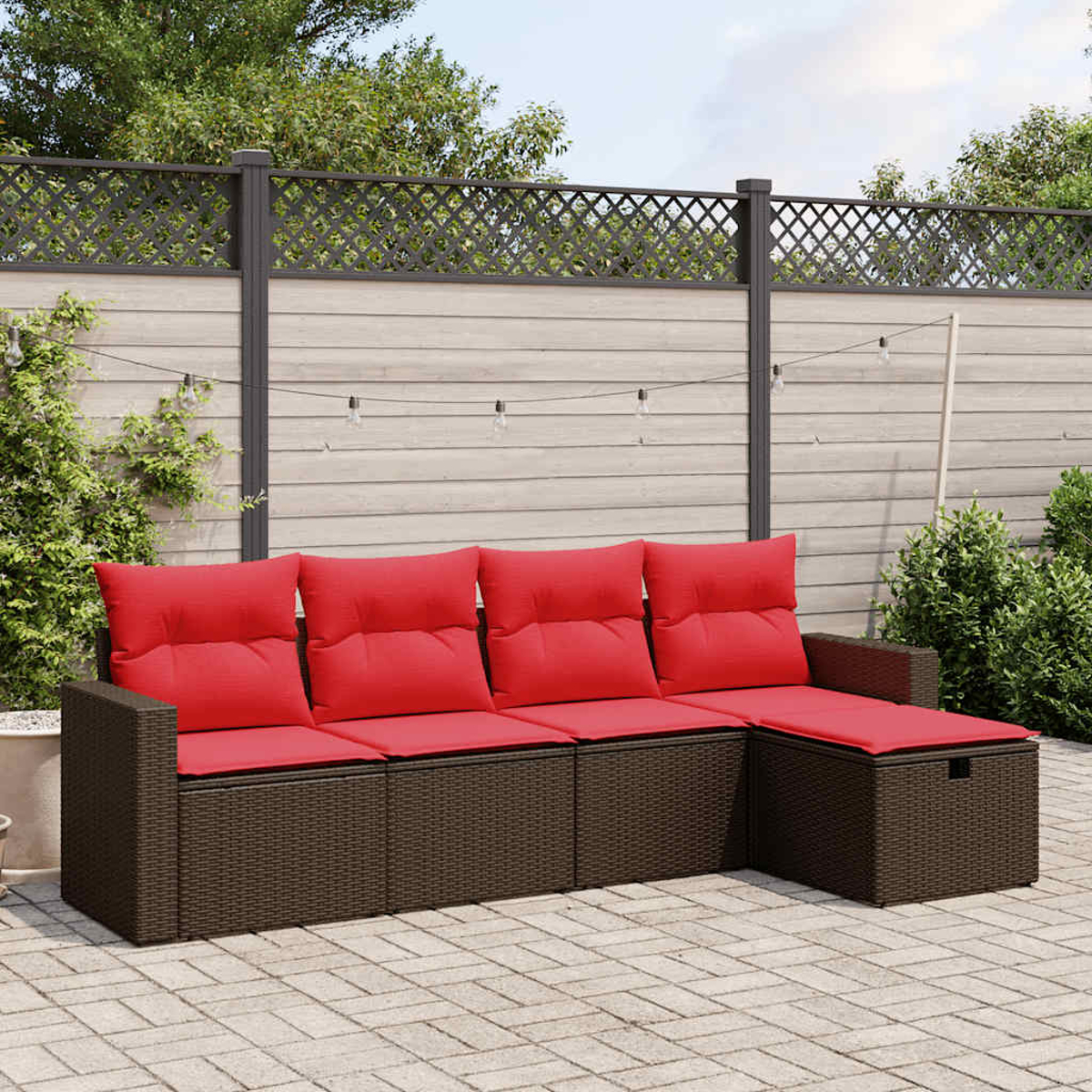 Un sofá seccional de mimbre marrón con cojines rojos sobre un patio pavimentado. Valla de madera y vegetación.