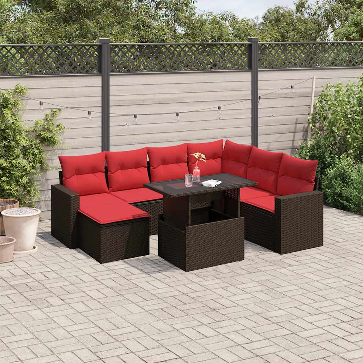Outdoor-Ecksofa mit roten Kissen und einem dunklen Tisch. Im Hintergrund ein Zaun und Pflanzen. Gepflasterte Terrasse.