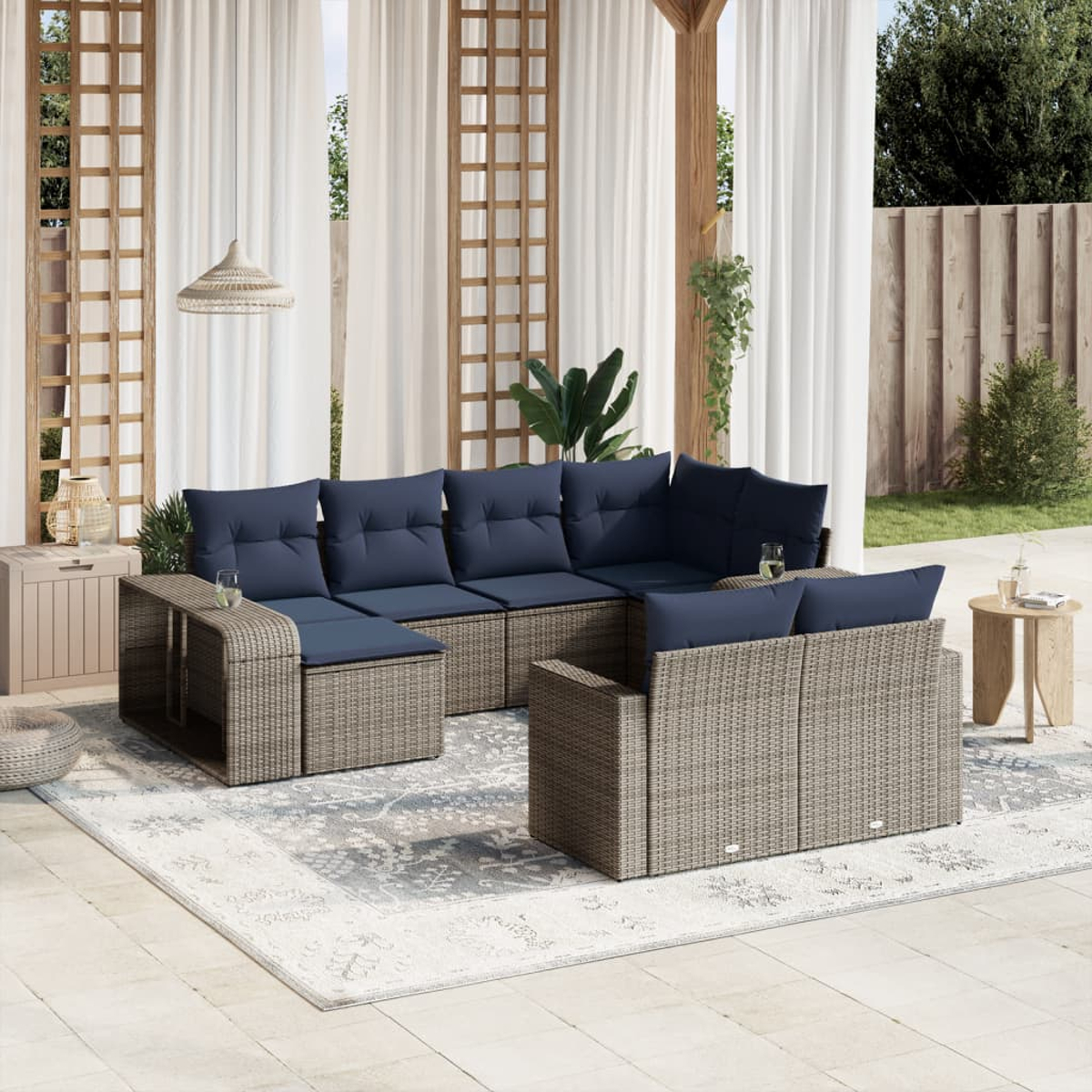 Zona de estar al aire libre con muebles de mimbre, cojines azul marino, una alfombra estampada y cortinas blancas. Fondo: valla de madera y vegetación.