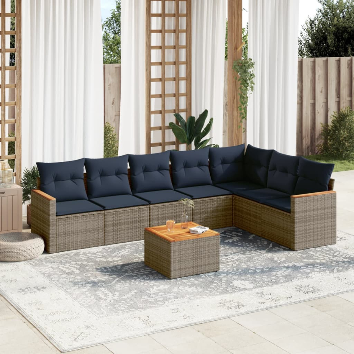 Ein Ecksofa mit blauen Kissen im Freien steht auf einem gemusterten Teppich. Davor steht ein kleiner Tisch. Weiße Vorhänge und ein Holzzaun sind sichtbar.