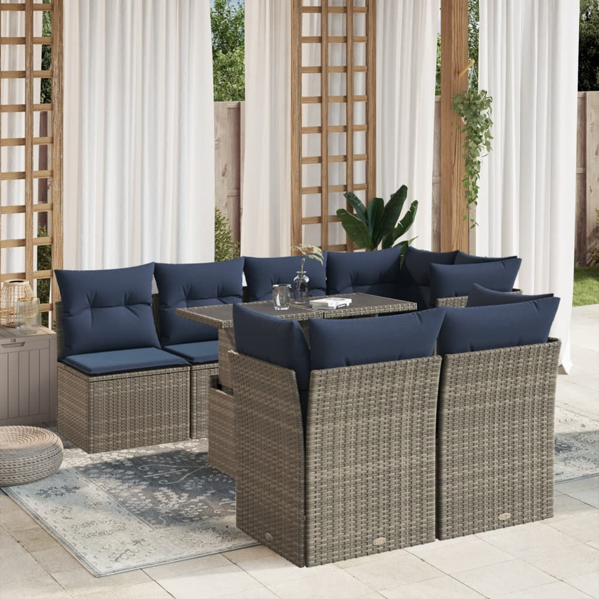 Juego de patio al aire libre con cojines azules, una mesa y cortinas. El juego está sobre una alfombra estampada. El fondo incluye vegetación y estructura de madera.