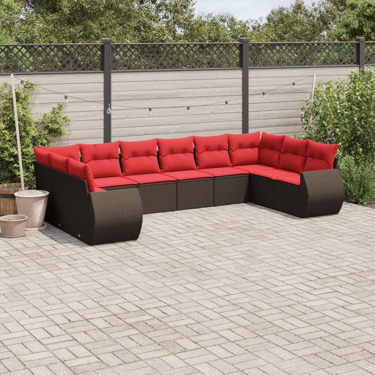Ein Ecksofa mit roten Kissen im Freien steht auf einer Terrasse. Dahinter befinden sich ein Zaun und Grün. Das Sofa ist braun.