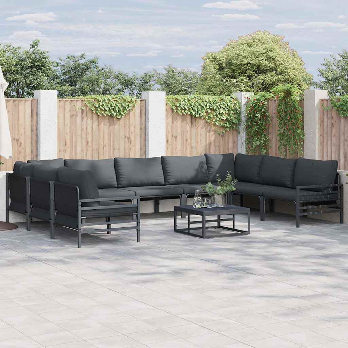 Outdoor-Ecksofa mit grauen Kissen. Ein kleiner schwarzer Tisch mit Pflanzen davor. Holzzaun und grünes Laub im Hintergrund.