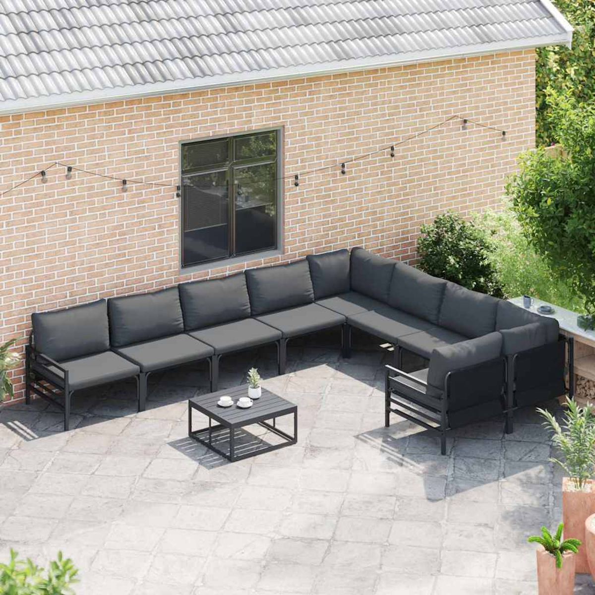 Eine Außenterrasse mit Ecksofa, Tisch und Pflanzen. Eine Backsteinmauer und ein Ziegeldach sind im Hintergrund zu sehen.