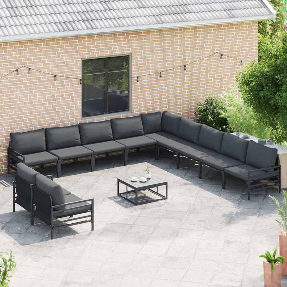 Außenbereich mit grauem Ecksofa, kleinem Tisch und zwei Sesseln auf einer Steinterrasse. Hintergrundmauer aus Ziegeln.