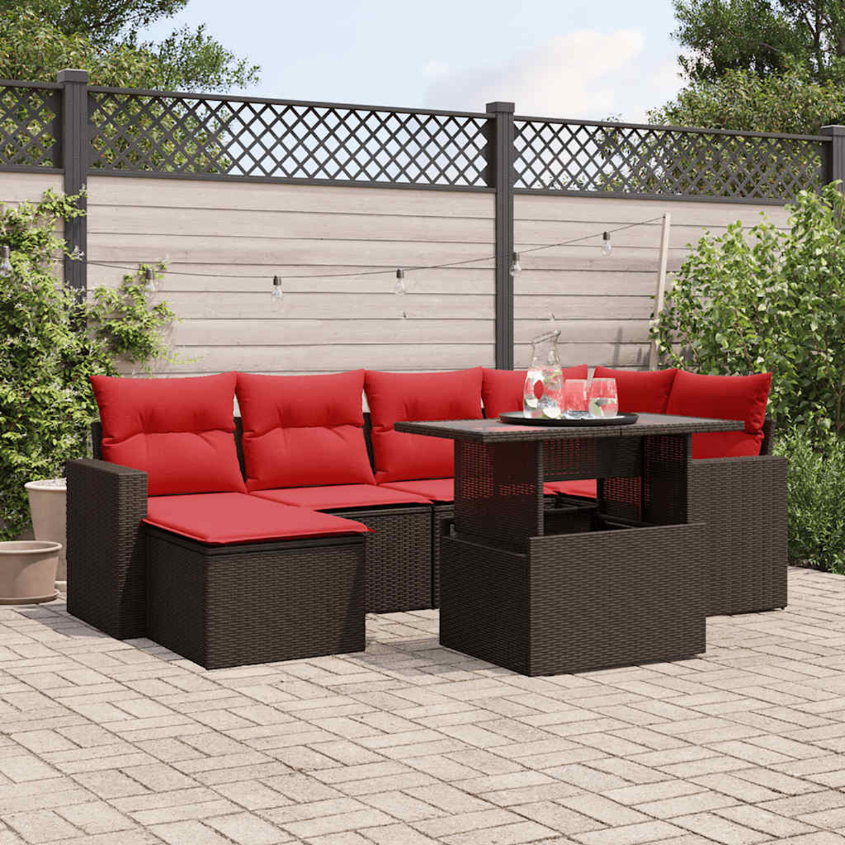 Gartenmöbel-Set. Braunes Korbmöbel, rote Kissen, Tisch mit Getränken und Gitterzaun im Hintergrund.