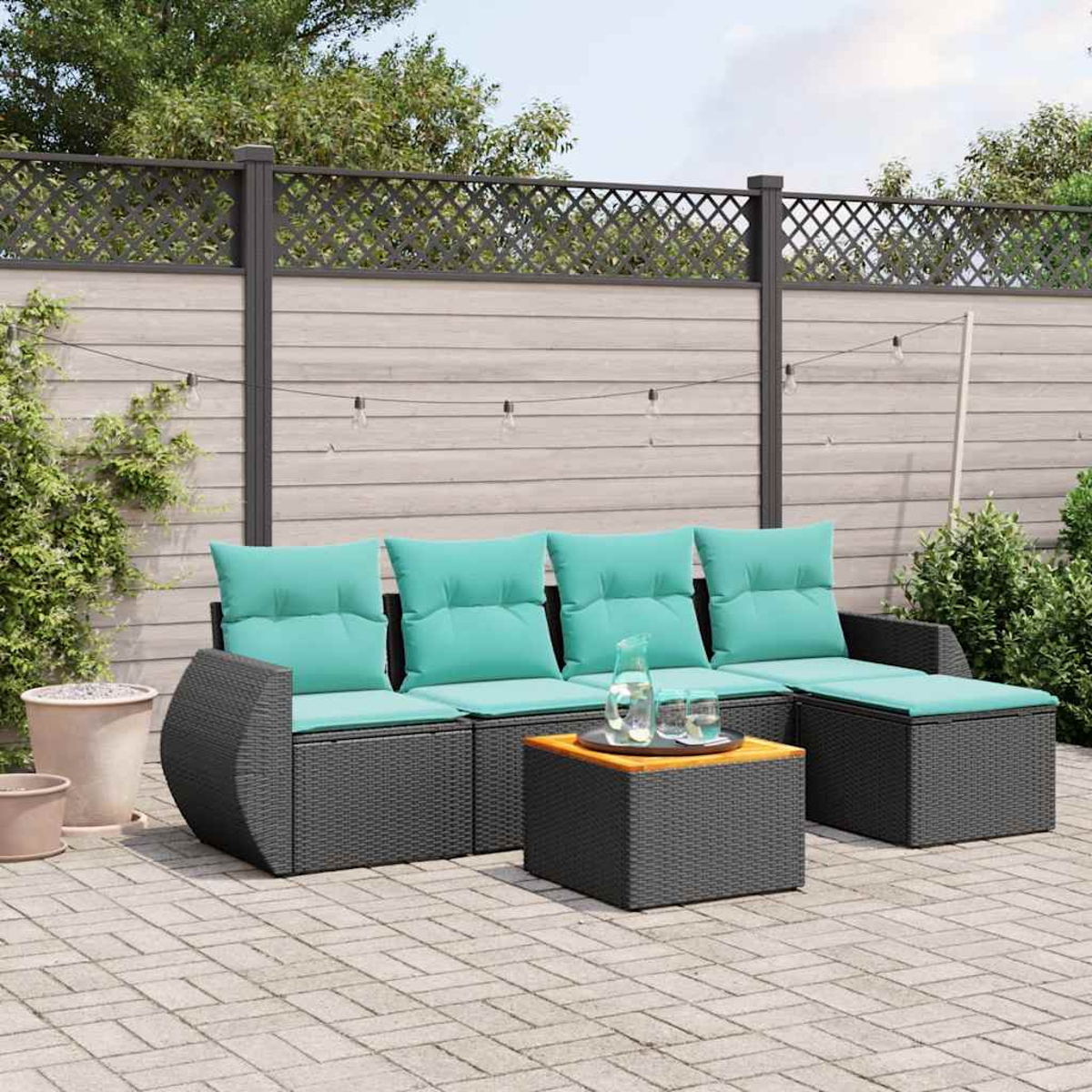 Juego de patio al aire libre con cojines verde azulado, muebles de mimbre grises y una mesa de madera. Una valla y plantas están en el fondo.