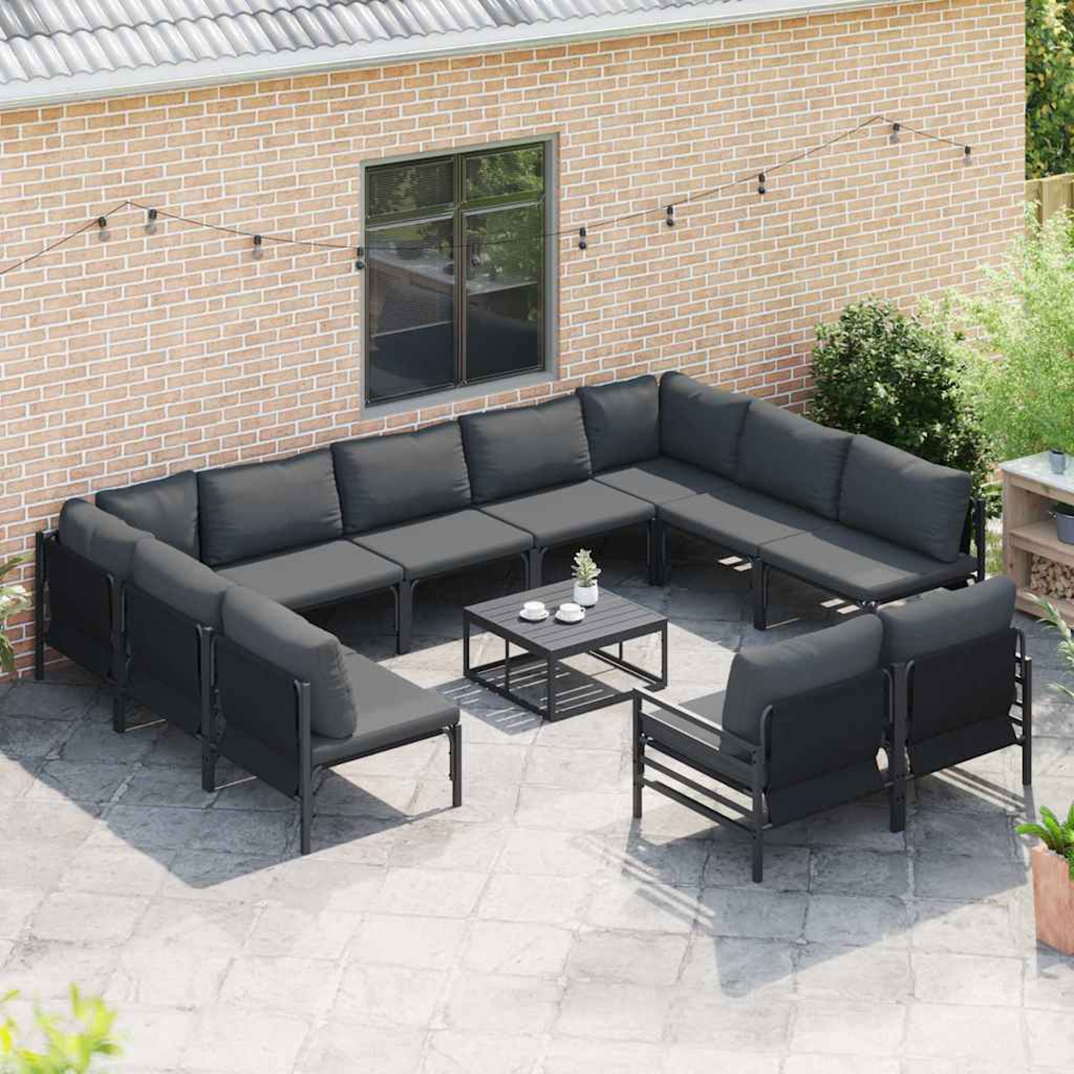 Outdoor-Ecksofa mit grauen Kissen und schwarzen Rahmen. Ein kleiner Tisch mit Tassen und Pflanzen. Backsteinmauer im Hintergrund.