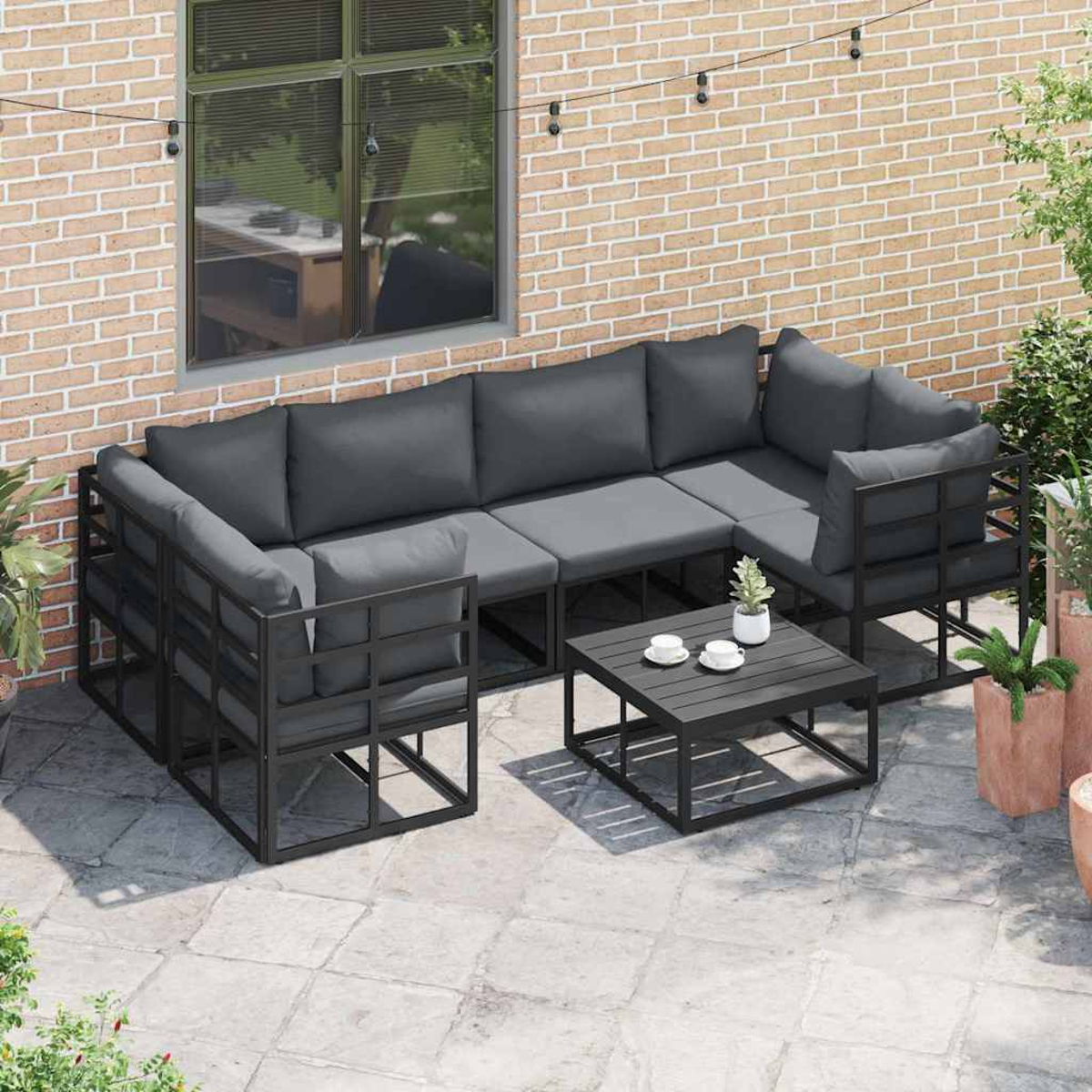 Outdoor-Ecksofa mit grauen Kissen und Couchtisch. Auf einer Terrasse in der Nähe einer Backsteinmauer und Pflanzen.